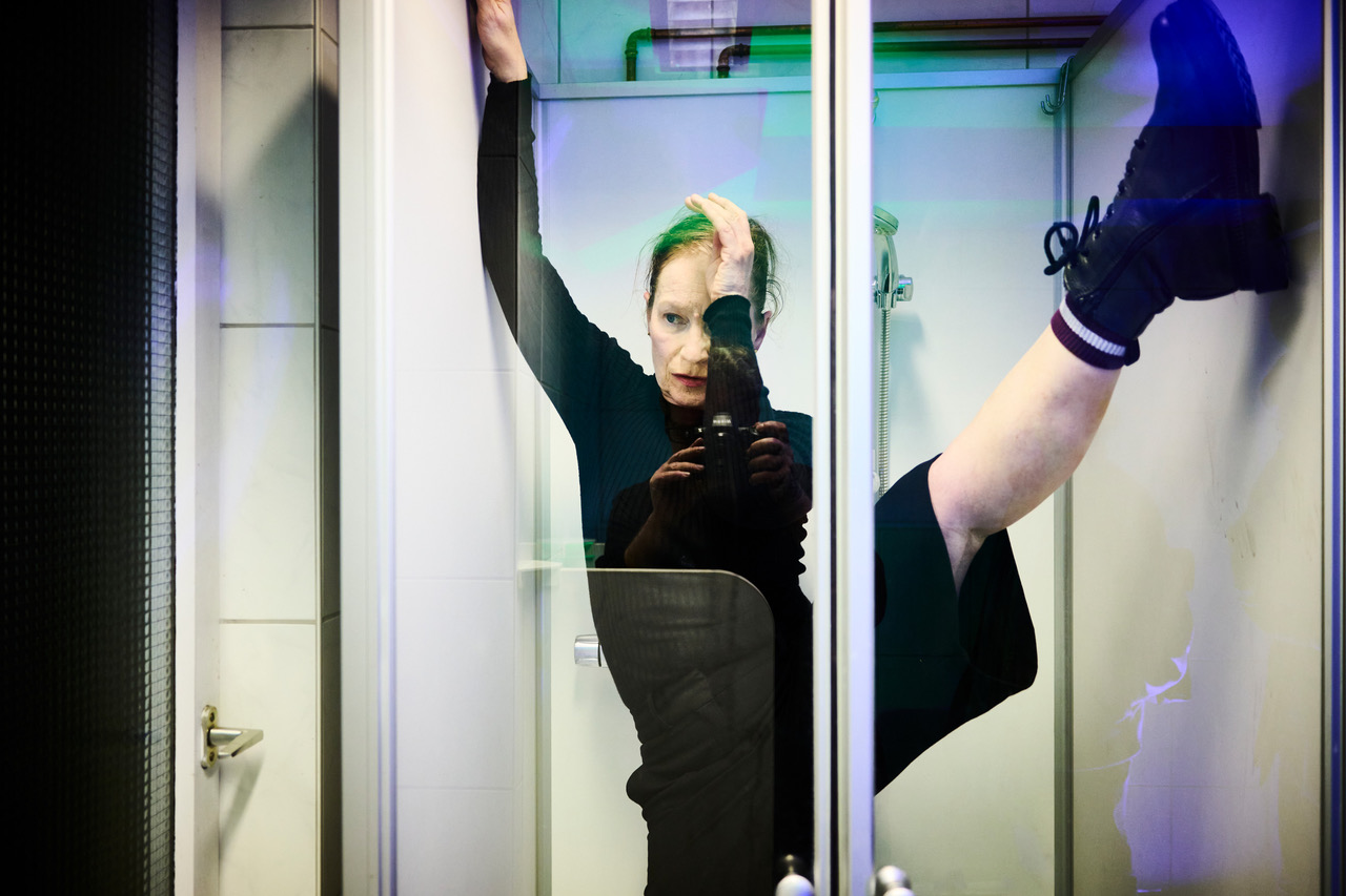 © Claudia Senoner Eine Tänzerin steht in einem schwarzen Kostüm mit einem hochgestreckten Beim in einer Duschkabine