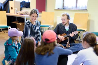 © Oliver Borchert Eine Musikerin und ein Musiker mit Gitarre sitzen mit Kindern im Kreis